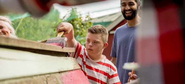 Kind mit Behinderung und junger Mann streichen Holzbauprojekt