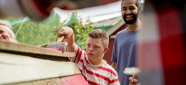 Kind mit Behinderung und junger Mann streichen Holzbauprojekt