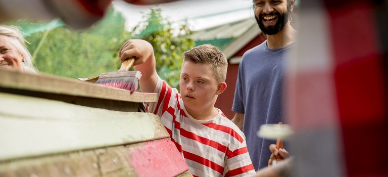 Kind mit Behinderung und junger Mann streichen Holzbauprojekt