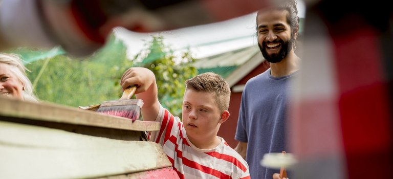 Kind mit Behinderung und junger Mann streichen Holzbauprojekt